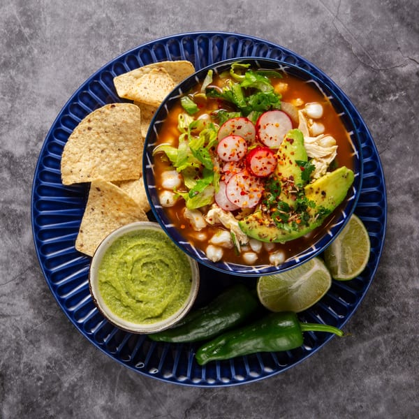 A bowl of Albertano's Mexican pozole with radishes, avocado, and lettuce, served with chips and guacamole.