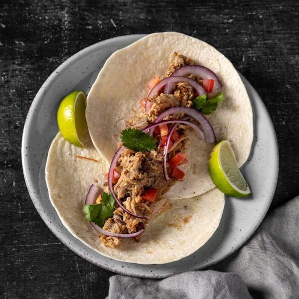 Two Albertano's carnitas tacos with red onion, tomato, cilantro, and lime wedges on a plate.