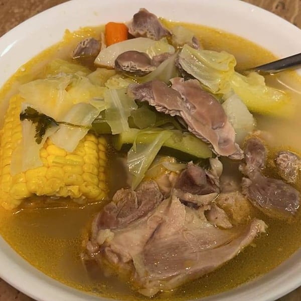 Close-up of a hearty Mexican soup with chicken, corn on the cob, cabbage, and carrots in a white bowl.